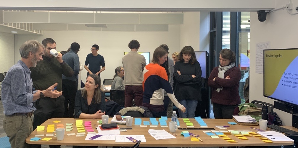 A group of people from the University of Exeter participating in a collaborative Matter of Focus research impact training workshop. Some are standing around a table covered with colorful sticky notes, papers, and cups, engaging in discussions. Others are standing in small groups or pairs in the background. A screen displays text reading 'Review in pairs' in the corner.