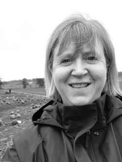 Black and white photo of Jennifer Wallace, smiling outdoors. The background features an open field with a distant tree line.