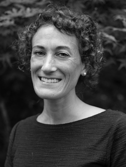 Black and white portrait of Kaitlynn Saldanha, smiling and looking at the camera. She has curly hair and is wearing a dark top. The background features soft-focus foliage.